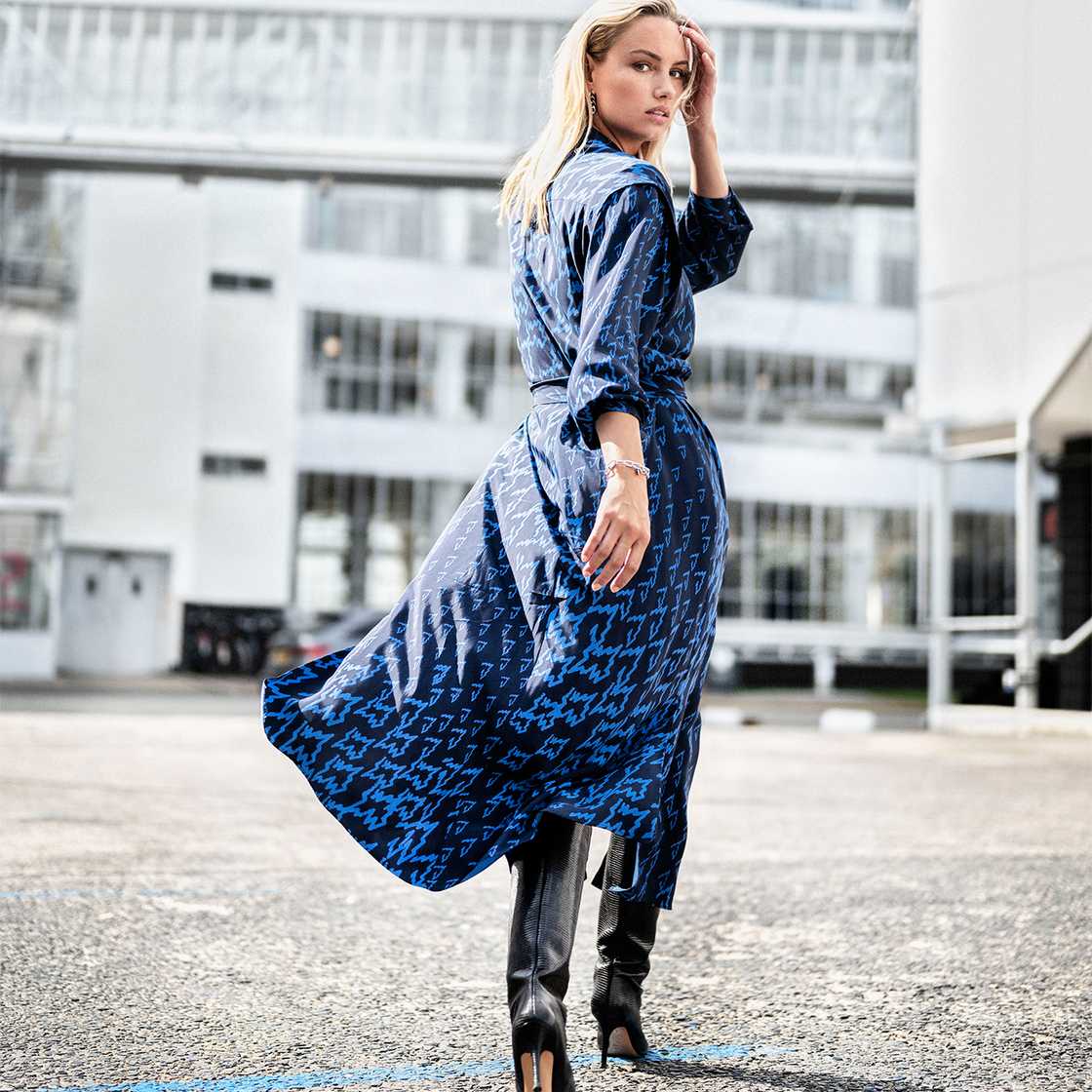 Image of a blond woman walking towards a building. She is looking back at the camera, wearing a blue dress and black boots