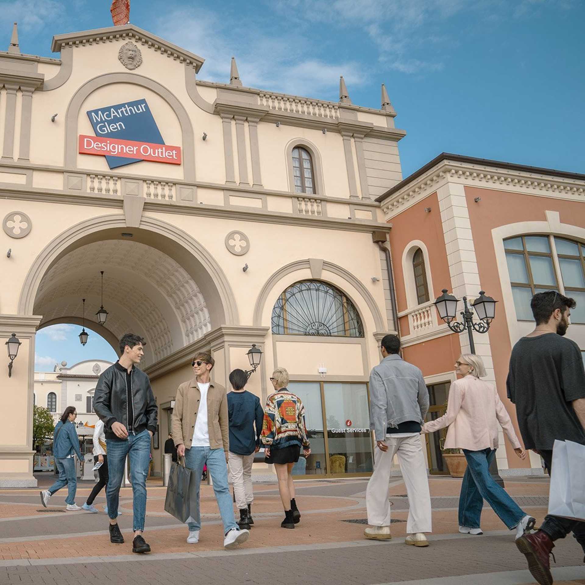 Group of people walking near the entrance of Noventa di Piave Designer Outlet, enjoying a sunny day of shopping.