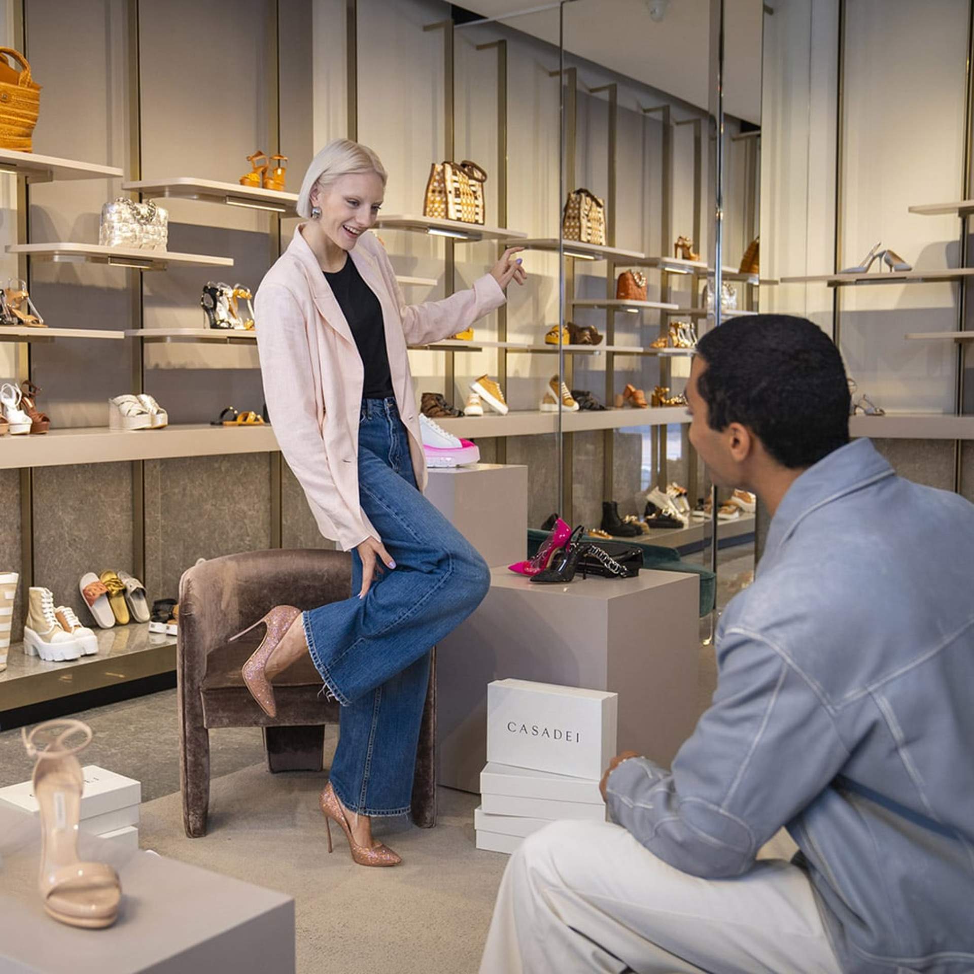 Woman trying on heels in a luxury boutique at Noventa di Piave Designer Outlet, talking to a seated man.