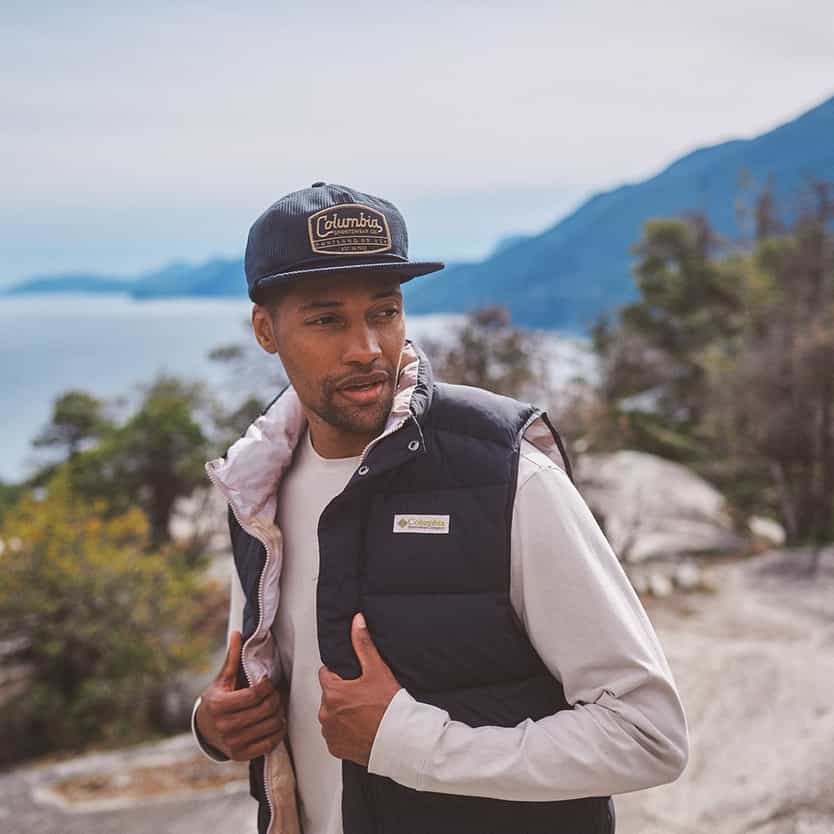 Columbia Man outdoors wearing a vest and cap with a scenic mountain and lake view.