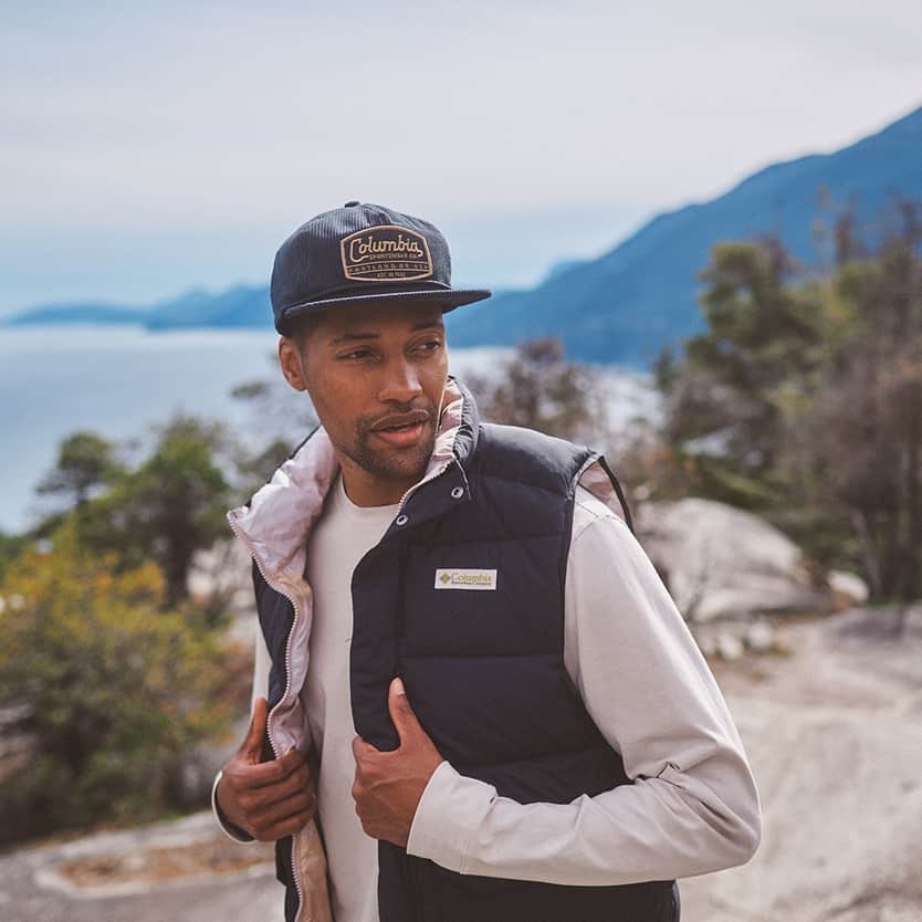 Columbia Man outdoors wearing a vest and cap with a scenic mountain and lake view.