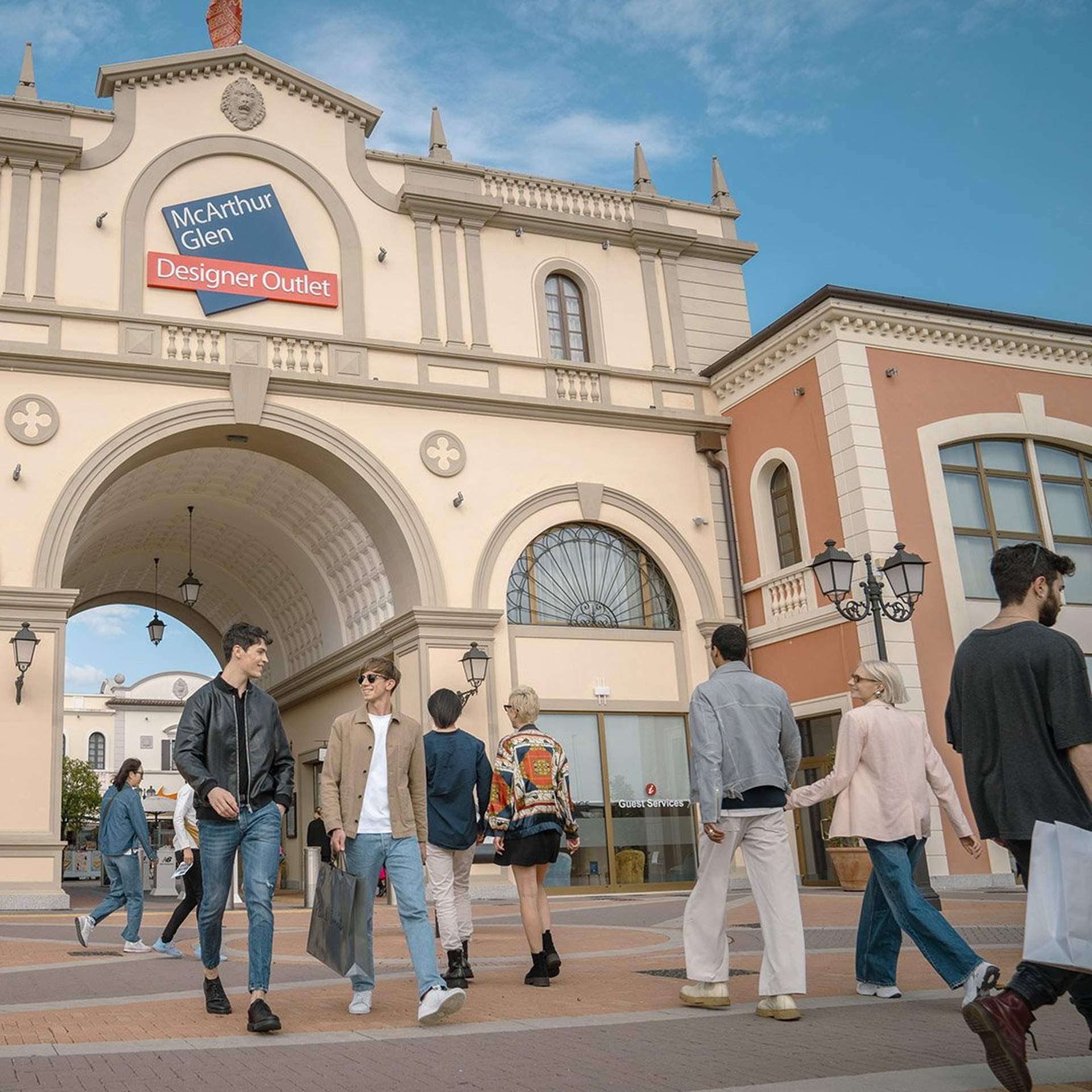 Group of people walking near the entrance of Noventa di Piave Designer Outlet, enjoying a sunny day of shopping.